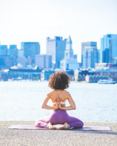Woman sitting on pier doing yoga, facing a city-scape